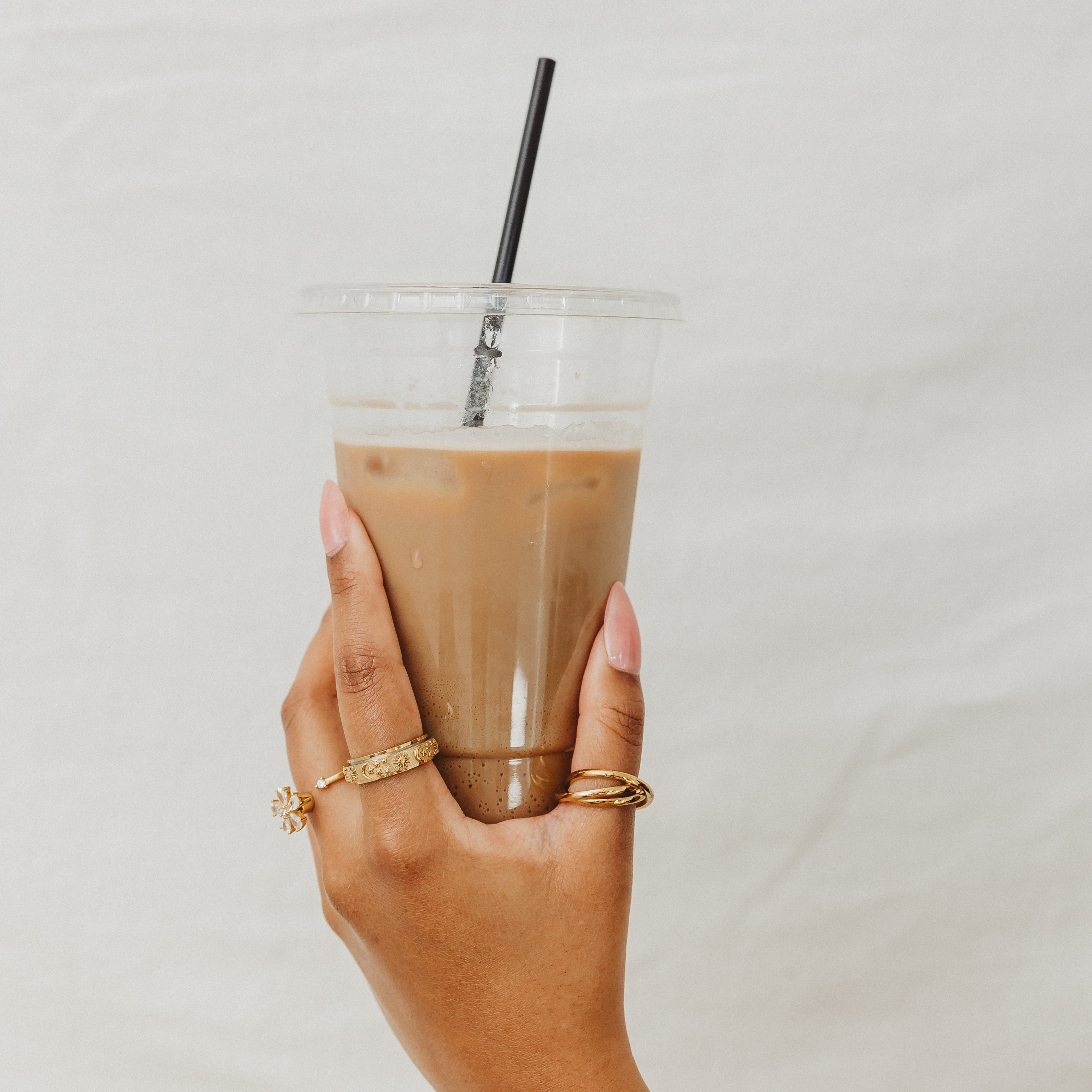 Hand holding a clear plastic cup with a iced coffee beverage and a straw against a plain background