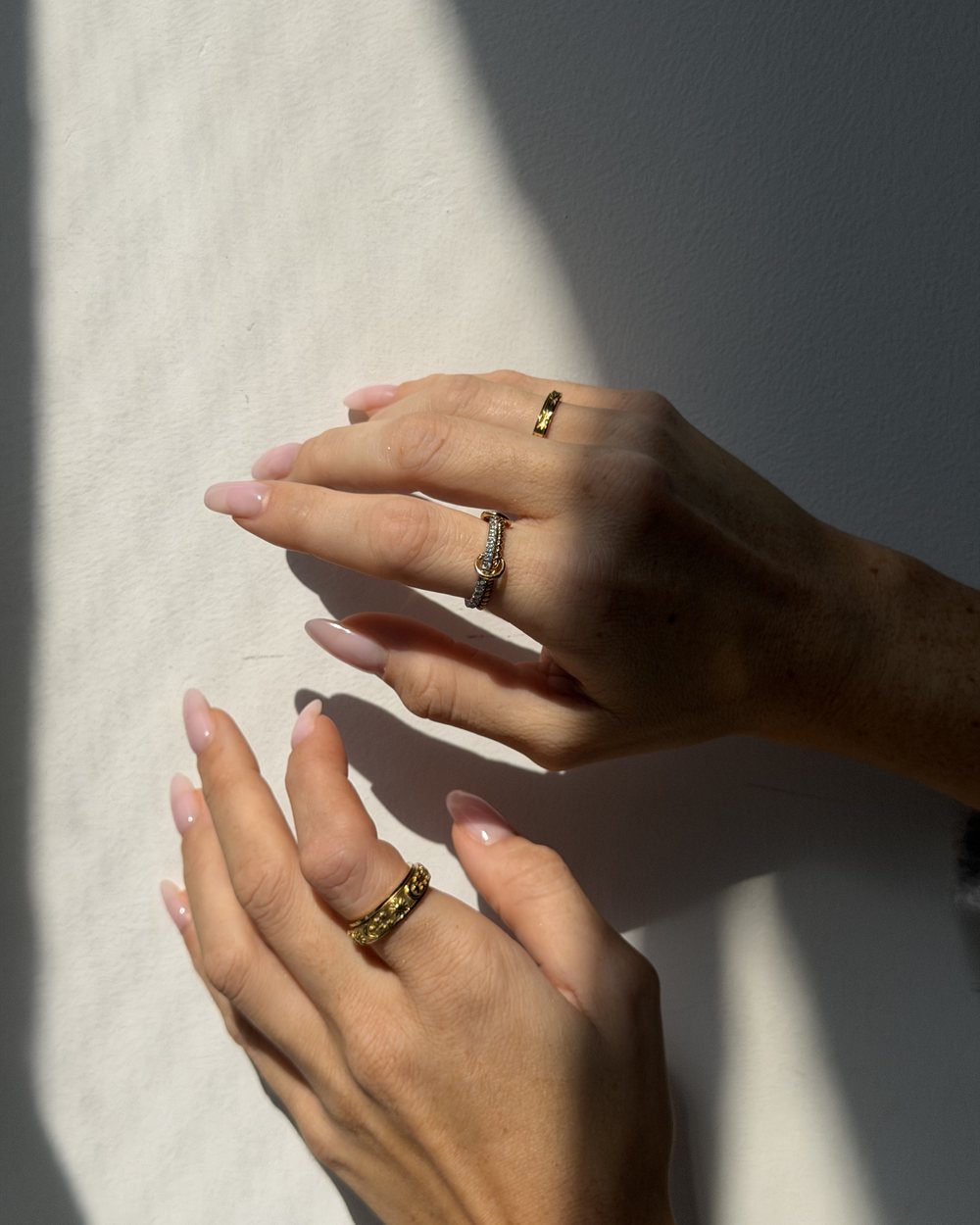 Two hands in the sun against a white wall, gold Anxi Rings on