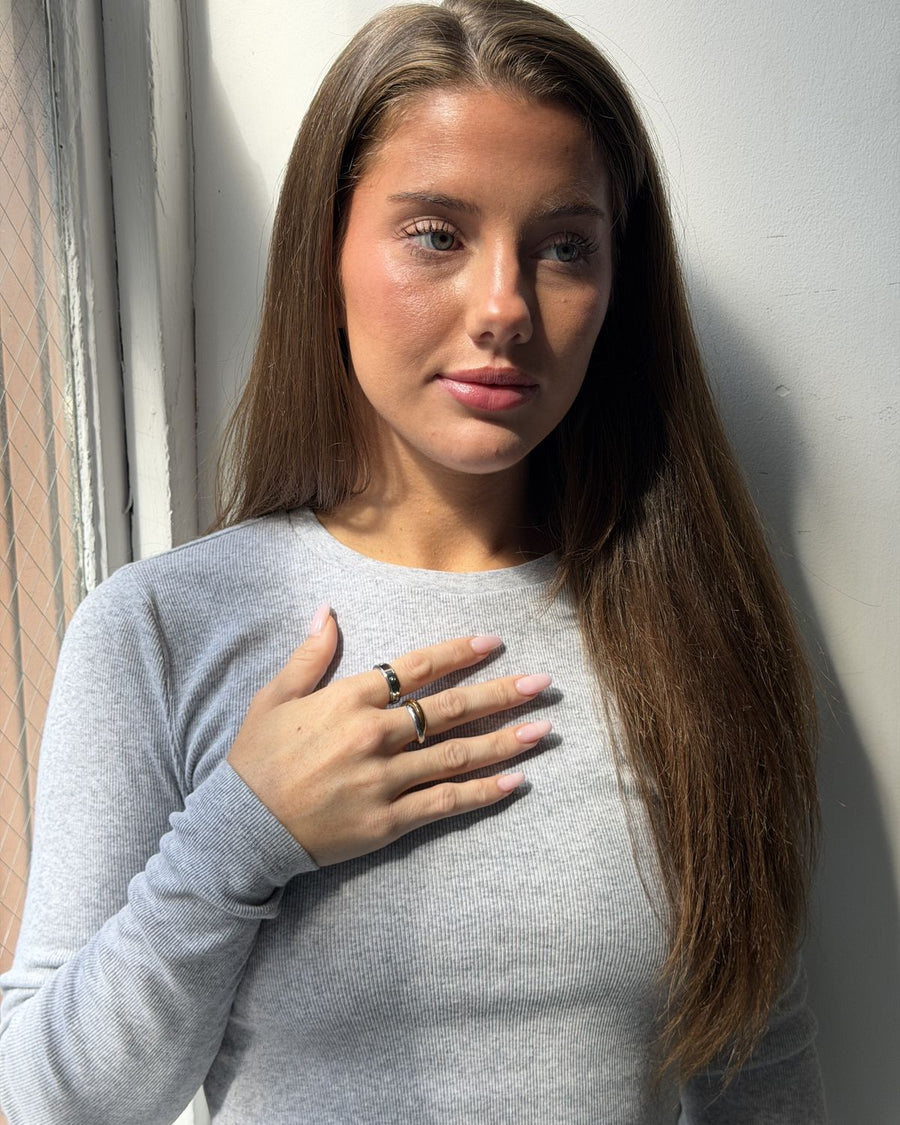 A woman in a grey top with her hand over her heart, wearing gold anxi rings