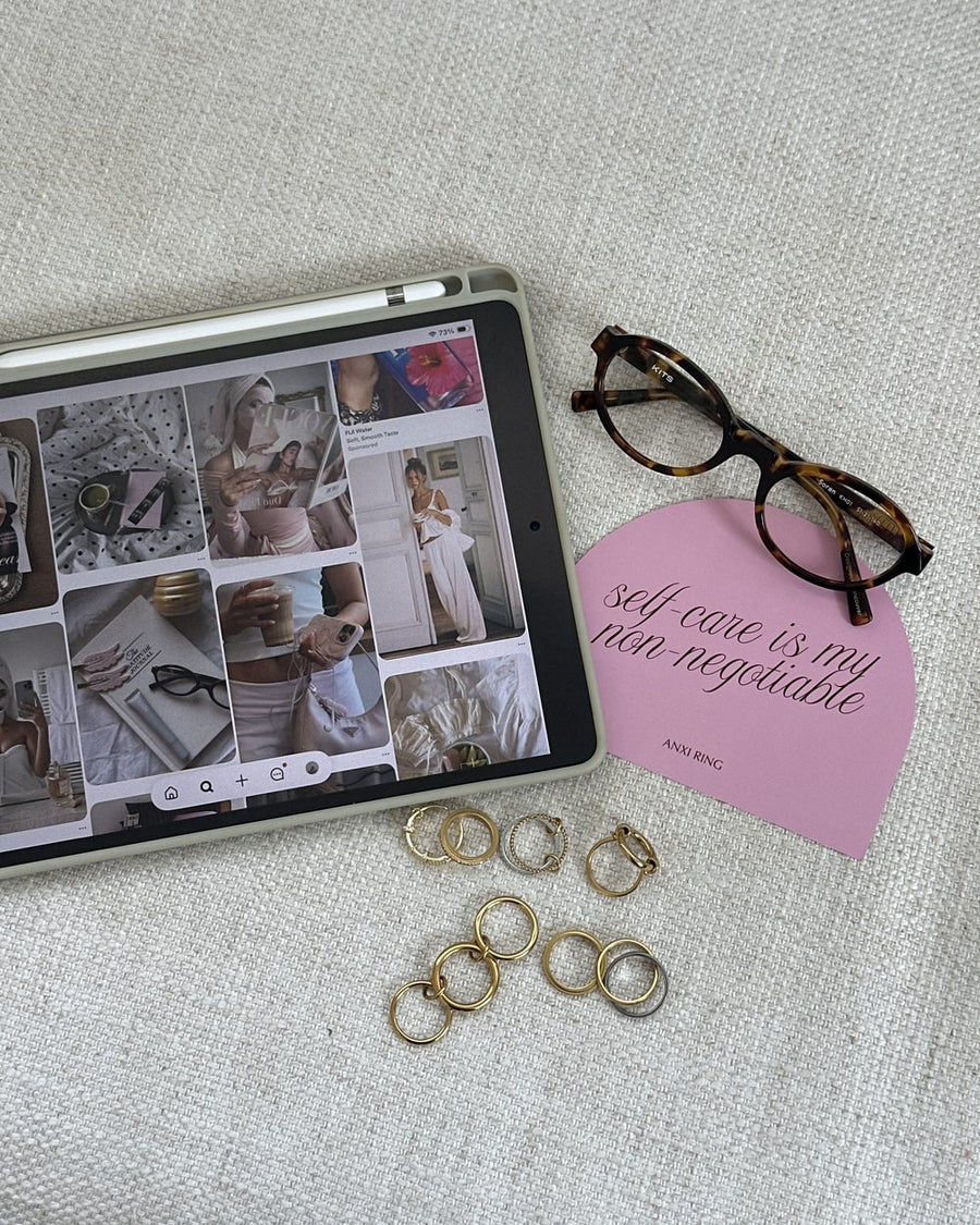 Gold Anxi Rings beside an iPad moodboard, tortoiseshell glasses and a pink self-care card