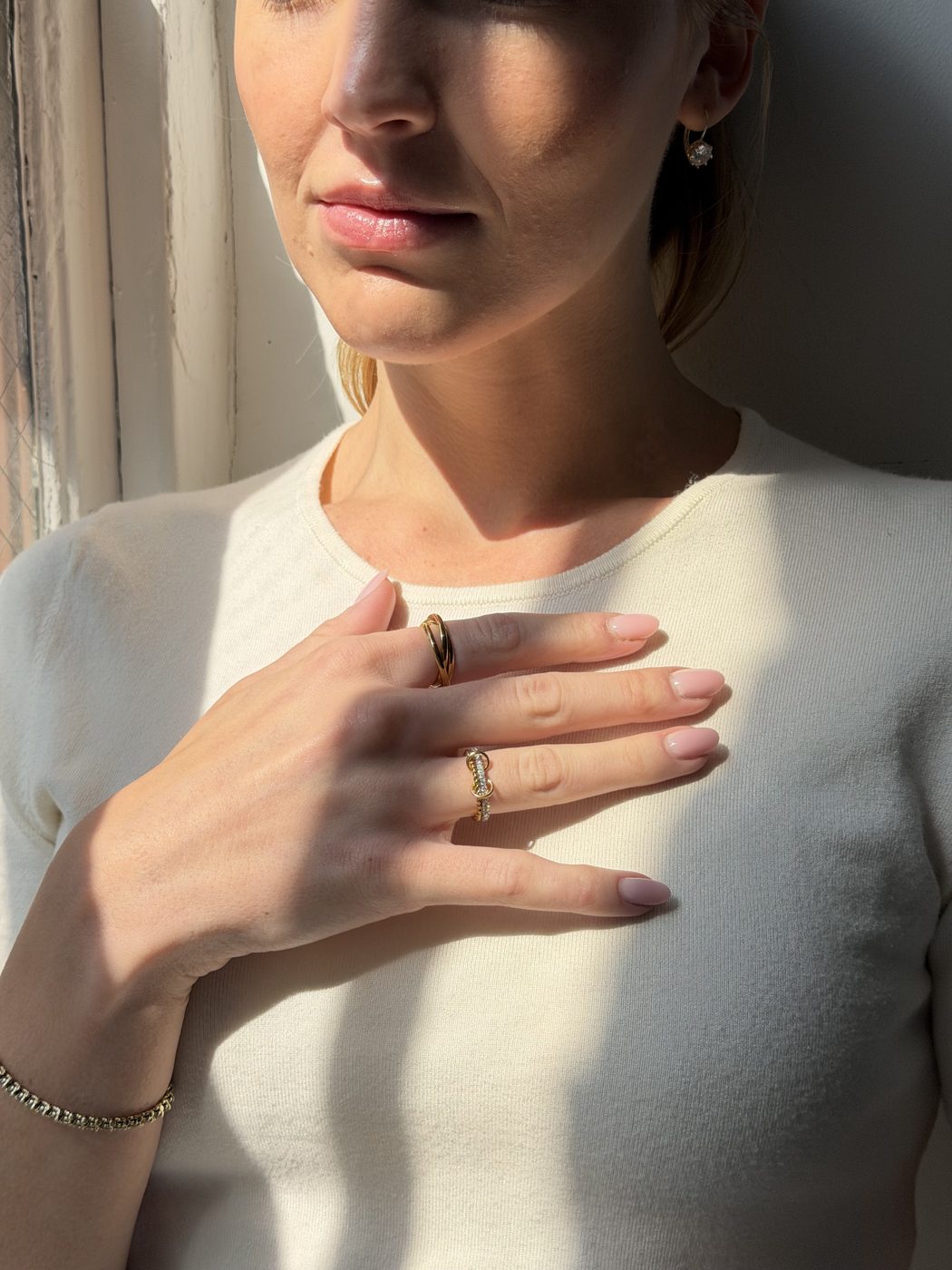 Hand resting on her chest in window light, wearing two gold Anxi Rings